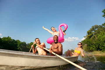 Happy group of friends having fun while laughting and swimming in river. Joyful men and women in swimsuit in a boat at riverside in sunny day. Summertime, friendship, resort, weekend concept.