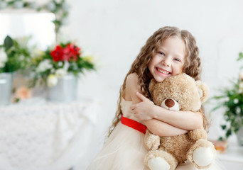 Portrait of expressive small beauty girl hugging huge plush bear, indoor shot on white room. Little girl with long hair playing with teddy bear. Place for text