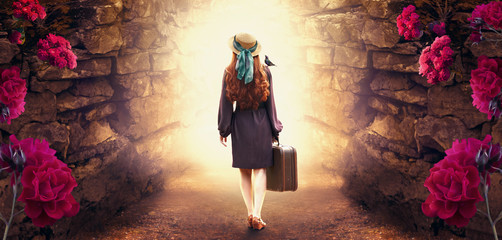 Young redhead lady woman in polka dot dress, hat and suitcase in retro style with starling bird on shoulder walking out through stone dungeon cave towards mystical glow. Idyllic tranquil fantasy scene