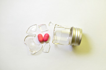 A broken small glass bottle with metal cap, two pink heart-shaped tablets fell out of it, on white background, top view