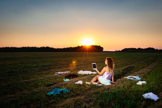 Woman sitting in the middle of a meadow with dropped unnecessary cloths and holding a tablet.  Environmental protection concept.