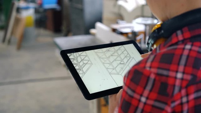 Female carpenter is using modern tablet in workshop looking at design of furniture on screen touching gadget. Technology, business and carpentry concept.