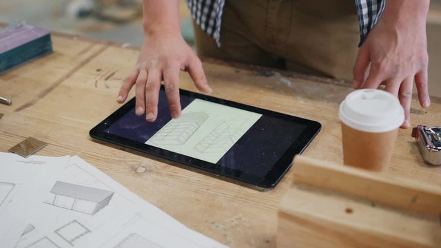 Close-up of male hand touching tablet screen in wood workshop indoors looking at furniture drawing planning construction. People, gadgets and business concept.