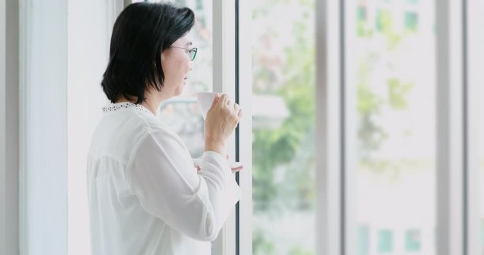 Smiling senior Asian woman standing and drinking coffee near large window.