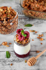 Layered breakfast with granola, yogurt and jam topped with raspberries in a glass jar on the white table
