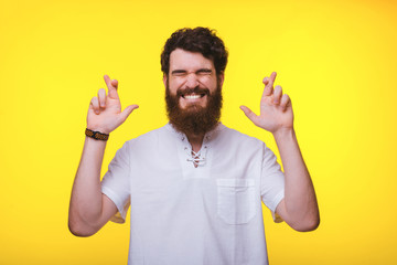 Young bearded man is standing with crossed fingers near yellow background.