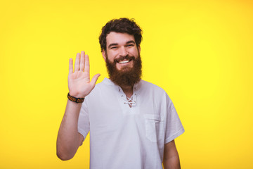 Smiling bearded man is waving at the camera near yellow wall.