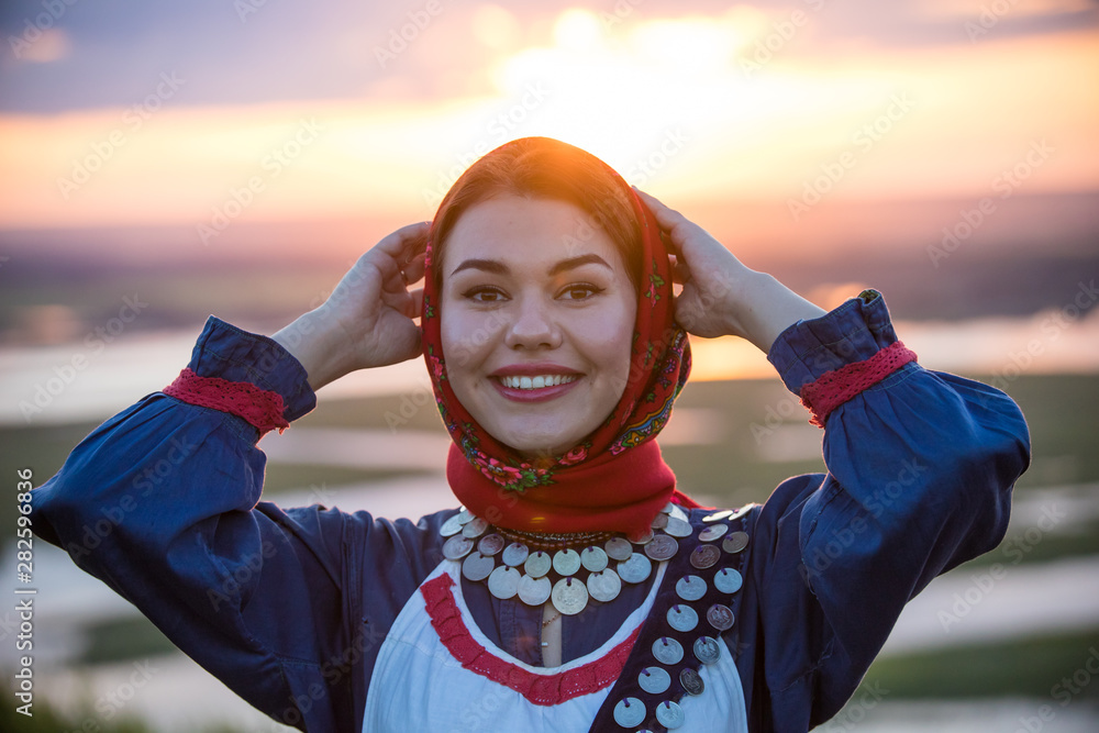 Wall mural Young smiling woman in traditional russian clothes on a background of the sunset