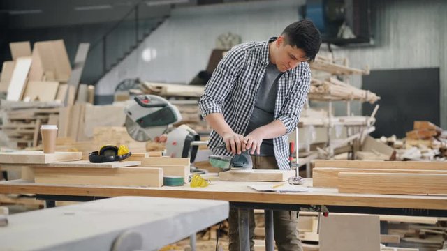 Good-looking carpenter is polishing piece of timber with electric machine in workroom standing at worktable holding equipment. Business and technology concept.