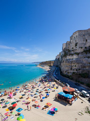 Dramatic setting of houses perched on cliff above beach with pale sand and blue sea, many bathers, Tropea, Italy - obrazy, fototapety, plakaty