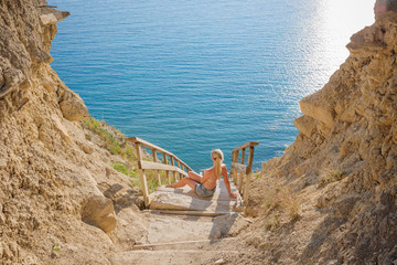 Summer lifestyle image of happy girl sitting on the cliff above the sea. Enjoying life and looking at the sea. Turquoise sea background