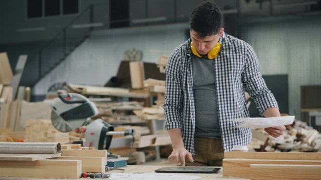 Man reading document working with tablet in wood workshop touching screen standing in workplace alone wearing casual clothing. People and devices concept.