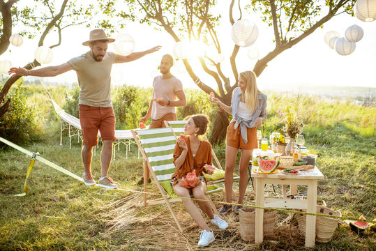 Young friends having fun, walking on a slackline during a picnic in the beautifully decorated garden on a sunset