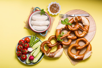 Pretzels, white bavarian sausages and vegetables on yellow background, german traditional food,...