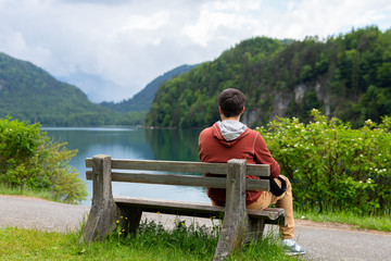 Young man sitting at bench and looking on Alpsee Lake surrounded by Alps mountains, Bavaria, Germany. Spring summer time. Blue clear water. Noone. Copy space. Space for text. Travel and Nature concept