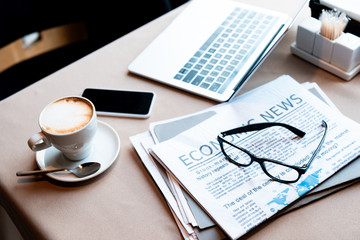 laptop and smartphone with blank screen, cup of coffee, documents, newspaper and glasses on table in cafe