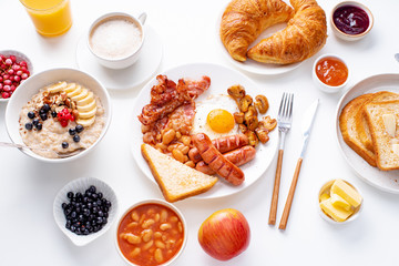 Top view flatlay with varieties of fresh breakfast: fried eggs with bacon and sausages, oatmeal with berries, fried toasts with jam and butter. White background.
