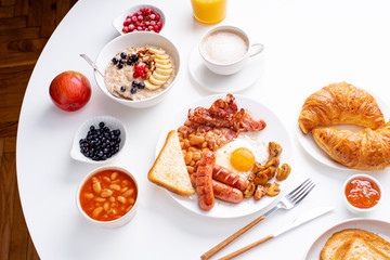 Top view flatlay with varieties of fresh breakfast: fried eggs with bacon and sausages, oatmeal with berries, fried toasts with jam and butter. White background.