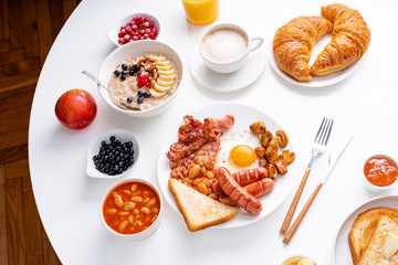 Top view flatlay with varieties of fresh breakfast: fried eggs with bacon and sausages, oatmeal with berries, fried toasts with jam and butter. White background.