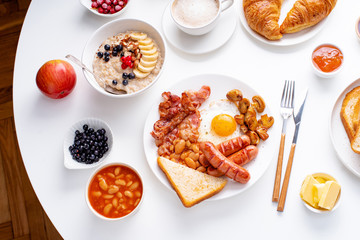 Top view flatlay with varieties of fresh breakfast: fried eggs with bacon and sausages, oatmeal with berries, fried toasts with jam and butter. White background.