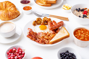 Top view flatlay with varieties of fresh breakfast: fried eggs with bacon and sausages, oatmeal with berries, fried toasts with jam and butter. White background.