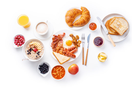 Top view flatlay with varieties of fresh breakfast: fried eggs with bacon and sausages, oatmeal with berries, fried toasts with jam and butter. White background.