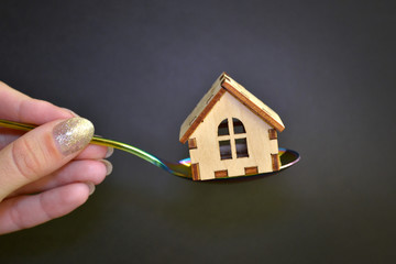 Wooden yellow toy house lies on rainbow metal spoon held by female hand on black background, the...