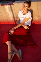 Bright summer lifestyle portrait of young pretty woman in eyewear, red skirt and white T-shirt, lying on a bright red floor outdoors.