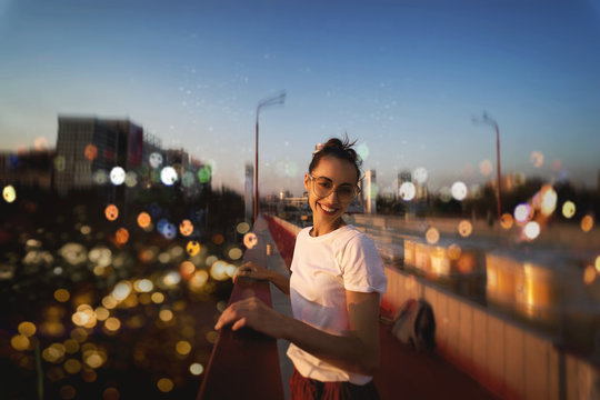 Bright summer lifestyle portrait of young pretty woman in eyewear, red skirt and white T-shirt, standing on the bright red bridge at evening with city background and blurred bokeh.