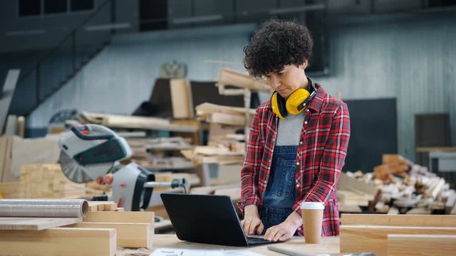 Female worker attractive young girl is using laptop in wood workshop typing standing in workplace alone focused on job. People, technology and industry concept.