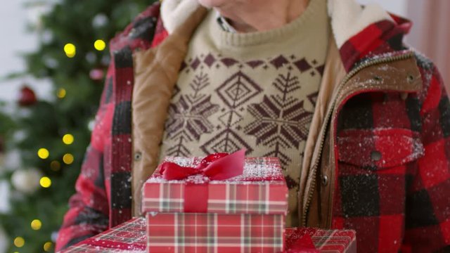 Tilt up portrait shot of elderly man wearing festive sweater and winter jacket covered in snowflakes standing in front of Christmas tree and holding stack of presents