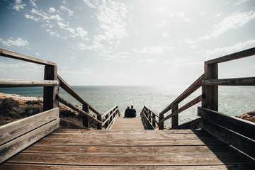 Couple in love sitting on stairs near ocean. Attractive pair enjoying ocean view on wooden path....