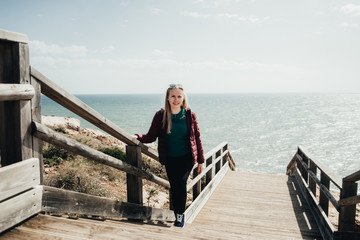 Blond woman standing on stairs near ocean. Attractive girl posing on wooden path. Female  enjoying...