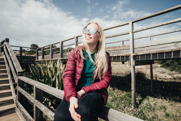Blond woman sitting on stairs near ocean. Attractive girl posing on wooden path. Female  enjoying...