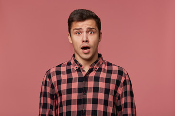 Fototapeta premium Young handsome amazed man in checkered shirt, looks at the camera with with wide open mouth in surprised expression, isolated over pink background.