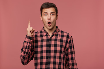 Young attractive wondered man wears in checkered shirt, looks at the camera with happy expression,...