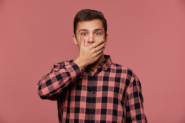 Fototapeta premium Portrait of young handsome shocked man in checkered shirt, looks at the camera with wide open eyes in surprised expression, covered mouth, hears unbelievable news, isolated over pink background.