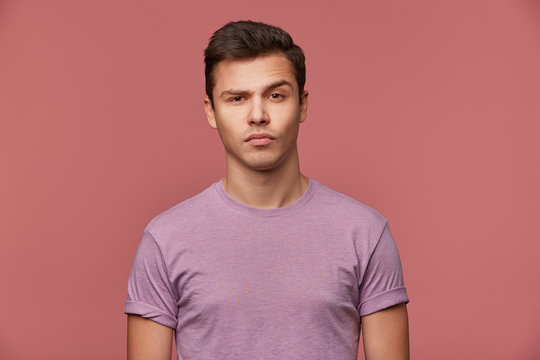 Portrait of handsome doubting young man wears in blank t-shirt, looks at the camera with raised eyebrow, stands over pink background.
