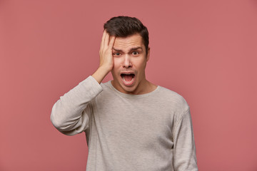 Obraz premium Young handsome shocked guy wears in checkered shirt, looks at the camera with wide open mouth in surprised expression, holds head, isolated over pink background.