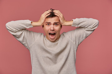 Young scared man in blank long sleeve, hears very bad news, holds head and looks at the camera with shocked expression, stands over pink background and screaming, looks afraid.