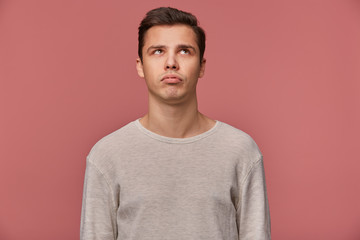 Young handsome thinking guy wears in checkered shirt, looks and away touches chin, looks bored,...