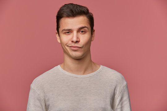 Portrait of handsome young man with raised eyebrow in disapproval, wears in blank t-shirt, looks at the camera with a grin and doubts, stands over pink background.