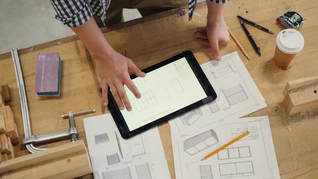Male hand touching tablet screen with technical drawings of furniture in workshop busy constructing handmade woodware. People, business and gadgets concept.