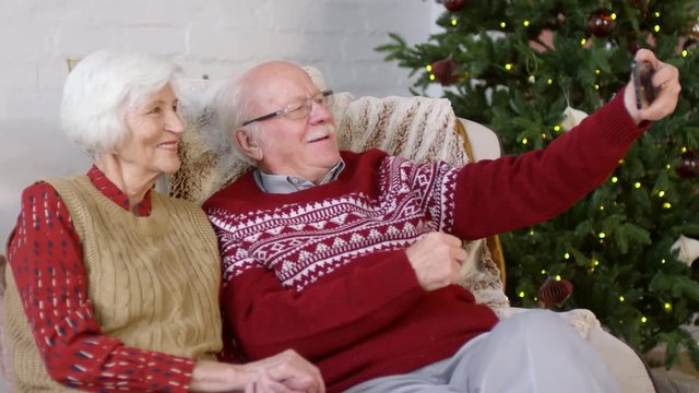 PAN shot of happy elderly couple in festive clothes sitting on sofa and taking selfie on mobile phone. Decorated Christmas tree in background