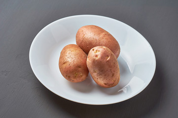 Three fresh raw potatoes on white ceramic plate on dark scratches concrete