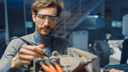 Professional Automotive Engineer in Glasses with a Computer and Inspection Tools is Testing an Used Electric Engine in a High Tech Laboratory with a Concept Car Chassis.