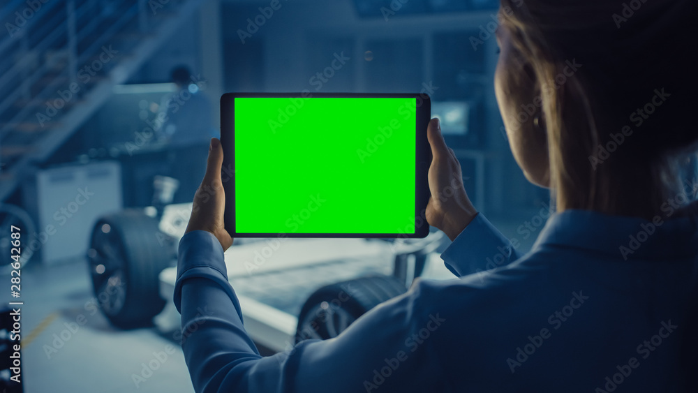 Poster Female Engineer Holds a Tablet Computer with Green Screen Mock Up and Looks at Electric Car Chassis Prototype with Wheels, Batteries and Engine Standing in a High Tech Development Laboratory.