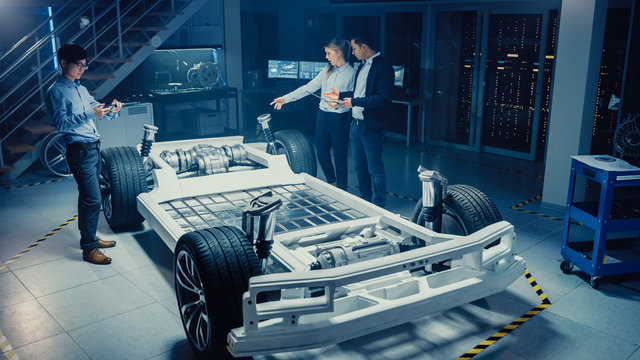 High Angle Shot of a Development Laboratory Room with Diverse Professional Team of Engineers and Scientists Working on a Electric Car Chassis with Wheels, Batteries, Engine and Suspension. 