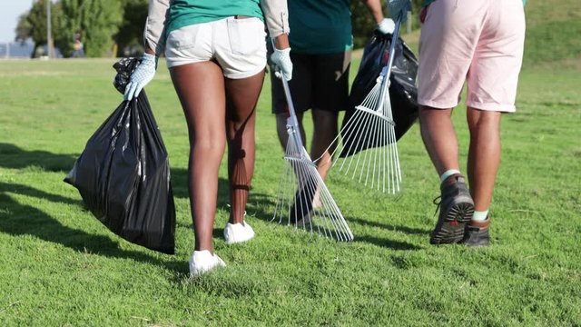Cropped shot of volunteers with garbage bags and racks. Low section of young people holding rakes and plastic bags and picking garbage in park. Recycling concept