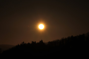 Beautiful orange ball on dark sky with shadows of trees. Sunset over black mountain. Eclipse of the sun in real time.